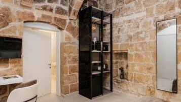 A stone-walled room with a white door, a black shelving unit, a mirror, and a white chair.