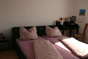 Een kamer met een bed, twee kussens, een nachtkastje, een stoel en een bureau met boeken en een agenda.