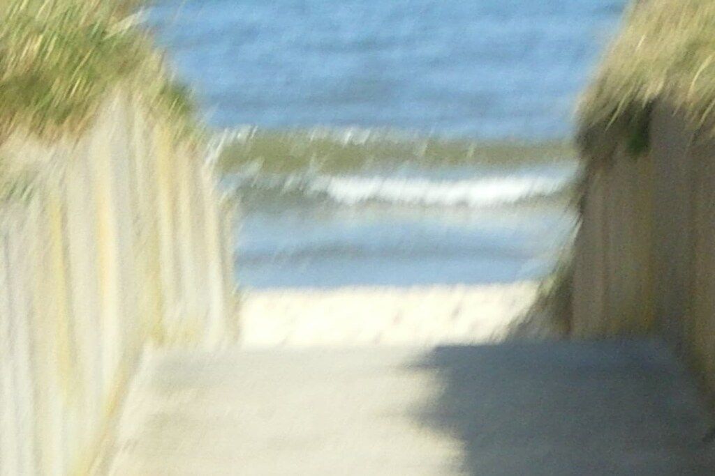 Caminho que leva a uma praia, com areia e ondas visíveis ao fundo.