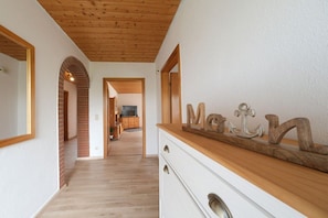 Ein Korridor mit Holzboden, einer weißen Wand und einer Holzkονstruktion an der Decke. Man sieht eine Holzkommode mit Schubladen und einen Spiegel.