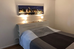 Ein ordentlich bezogenes Bett mit einer Streifenoptik-Decke, ein weißer Nachttisch mit zwei Lampen und ein gerahmtes Stadtbild über dem Bett.