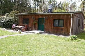 A wooden cabin with a green door, a small porch with a table and chairs, and a chimney. - Bungalow with 2 bedrooms (Reinhardtsdorf-Schöna)