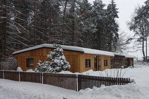A wooden cabin with a snow-covered roof, surrounded by a wooden fence and a snow-laden landscape. - Bungalow with 2 bedrooms (Reinhardtsdorf-Schöna)