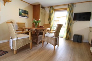 A dining area with a wooden table and wicker chairs, a vase with flowers, and a television on the wall. - Bungalow with 2 bedrooms (Reinhardtsdorf-Schöna)