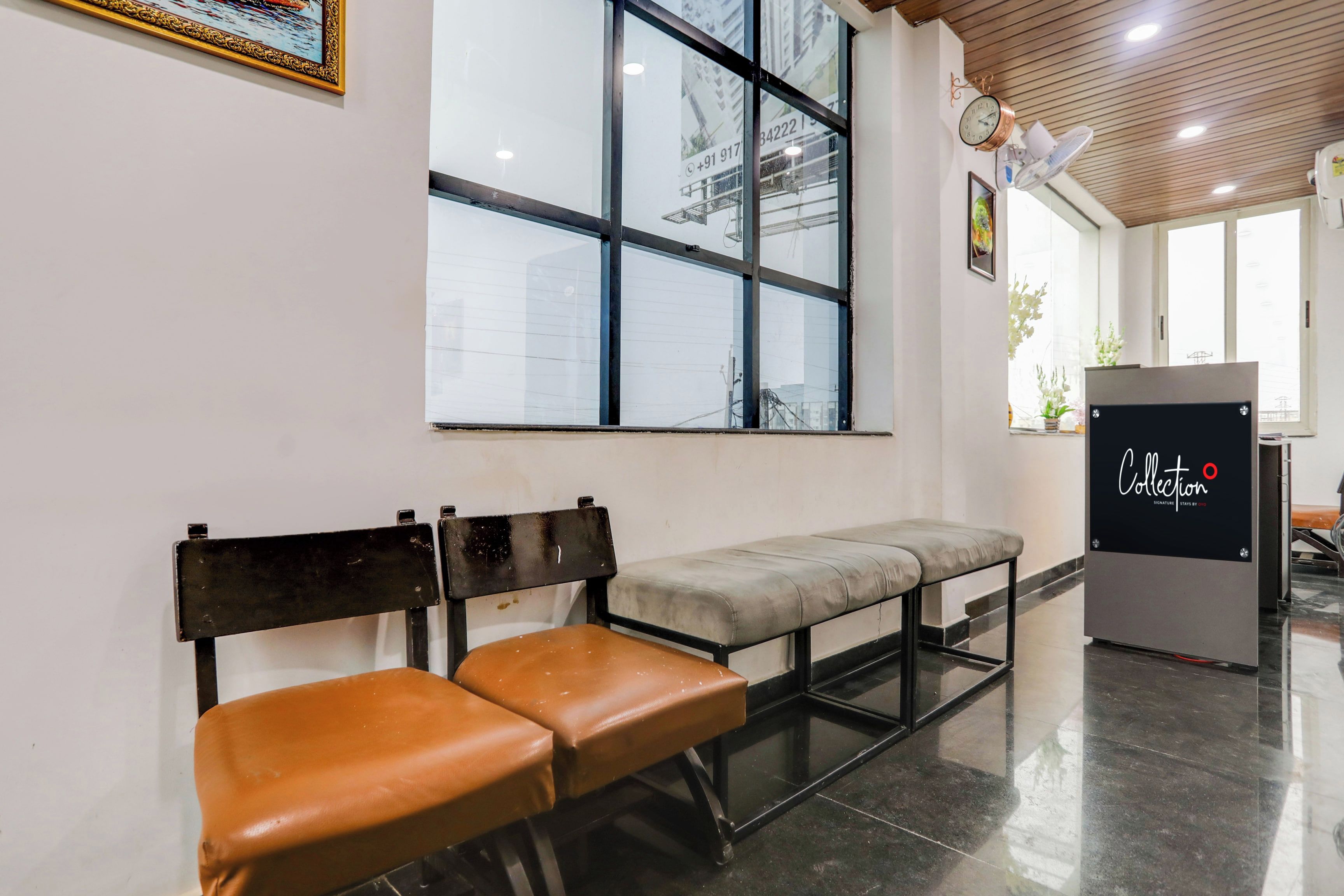 A waiting area with two wooden benches, a large window with a view of buildings, a clock, and a refrigerator with a logo.