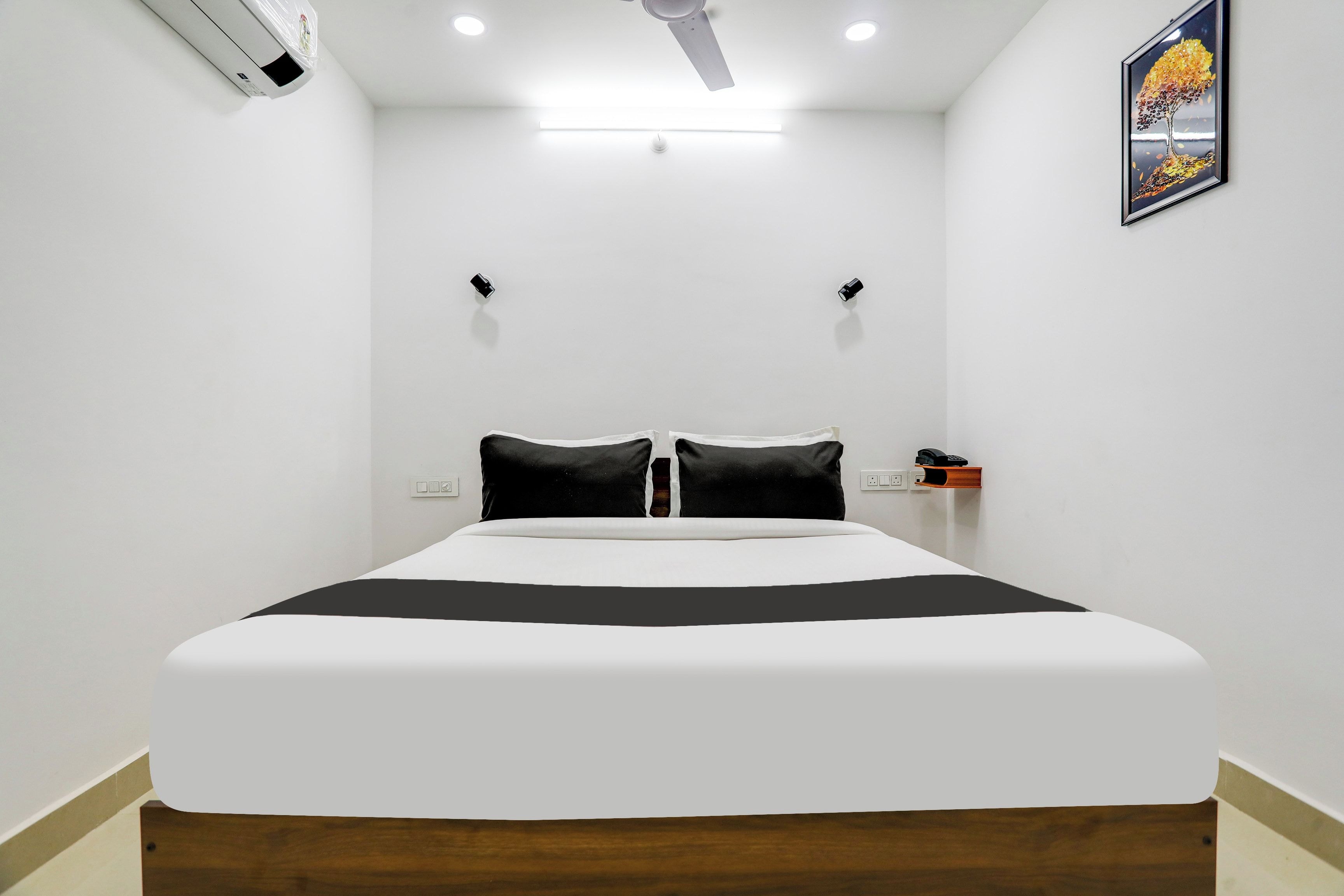 A bedroom with a bed, a ceiling fan, a wall-mounted air conditioner, a bedside table with a phone, and a framed picture on the wall.