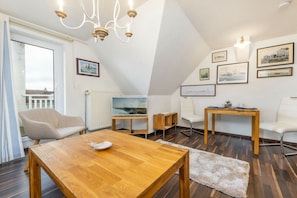 A modern living room with a wooden dining table, a white sofa, a white armchair, and a small wooden side table. There are framed pictures on the wall, a chandelier, and a view of the outside through a large window. - Holiday apartment in Binz with swimming pool (Binz)