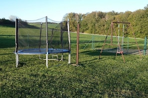 Ein Trampolin und eine Schaukel auf einer Wiese, im Hintergrund ein Zaun und Bäume.