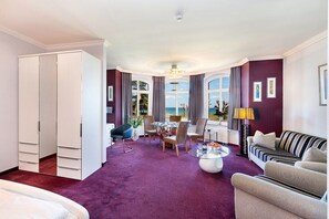 A hotel room with a bed, a seating area with a sofa and chairs, a dining table with chairs, and a large window with a view. - Bellevue in the Villa Glückspilz (Binz)