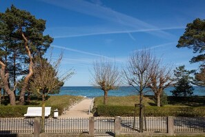 Een pad dat naar een strand leidt, met een bankje en een poort, omgeven door bomen en een heldere lucht.