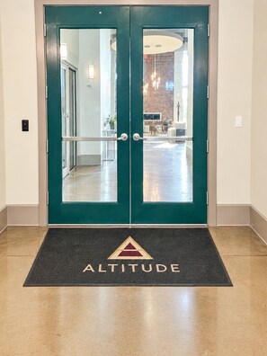 Ein Paar Doppeltüren mit einem Glasfenster, eine Fußmatte mit dem Wort 'ALTITUDE' und einem Logo sowie ein Flur, der zu einer Sitzecke führt.