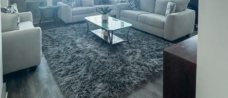 A modern living room with a large window, a grey shag rug, and a glass coffee table.