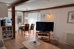 Een woonkamer met een televisie, een eethoek met stoelen, een boekenkast, en een keuken in de achtergrond.