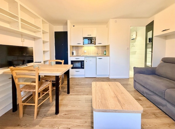 Un espacio habitable moderno con comedor, sofá gris, televisión y cocina con muebles y electrodomésticos blancos.