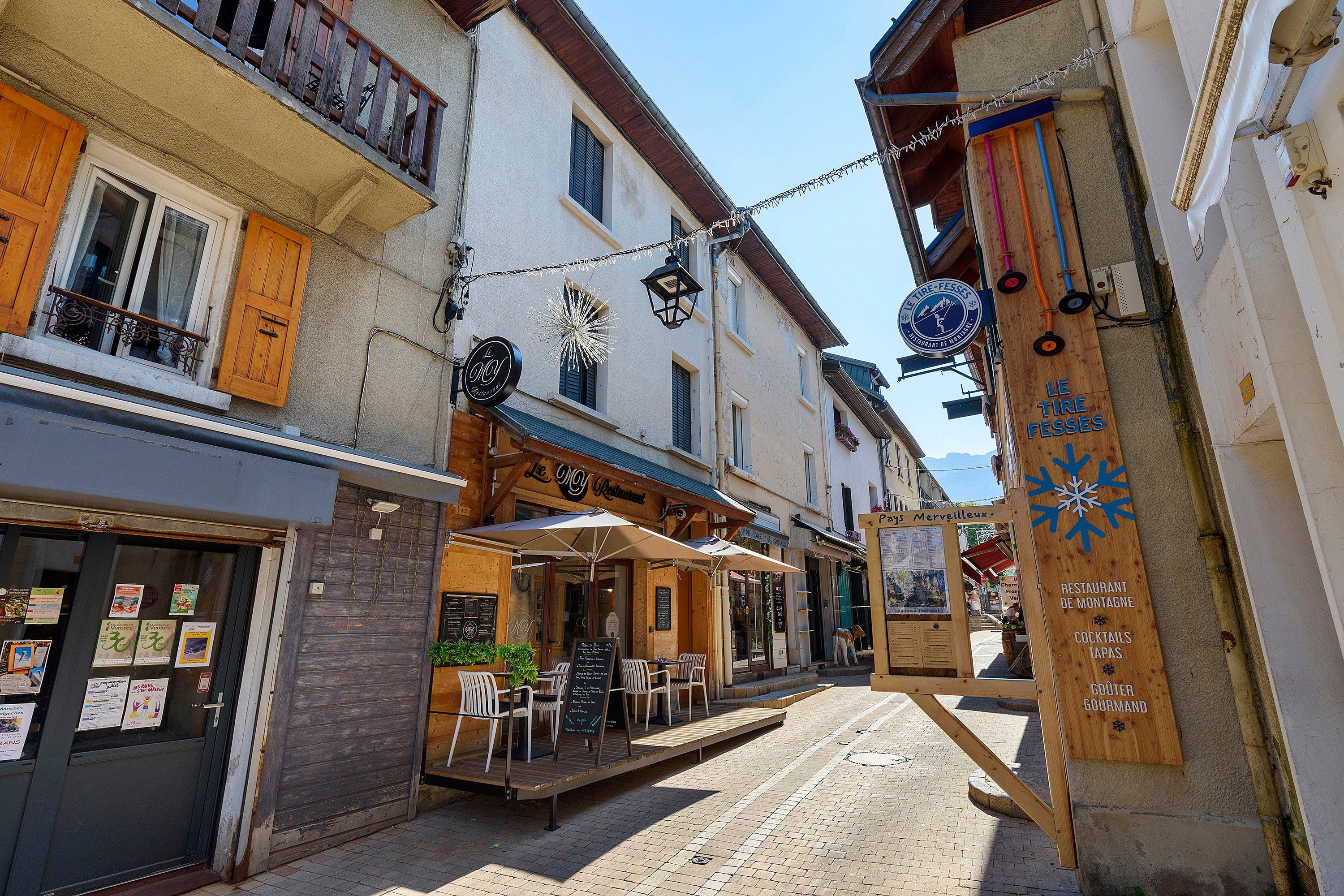Une rue étroite bordée de bâtiments des deux côtés, avec des tables en terrasse pour un restaurant, et une enseigne indiquant un restaurant appelé « Le Tire-Fesses ».