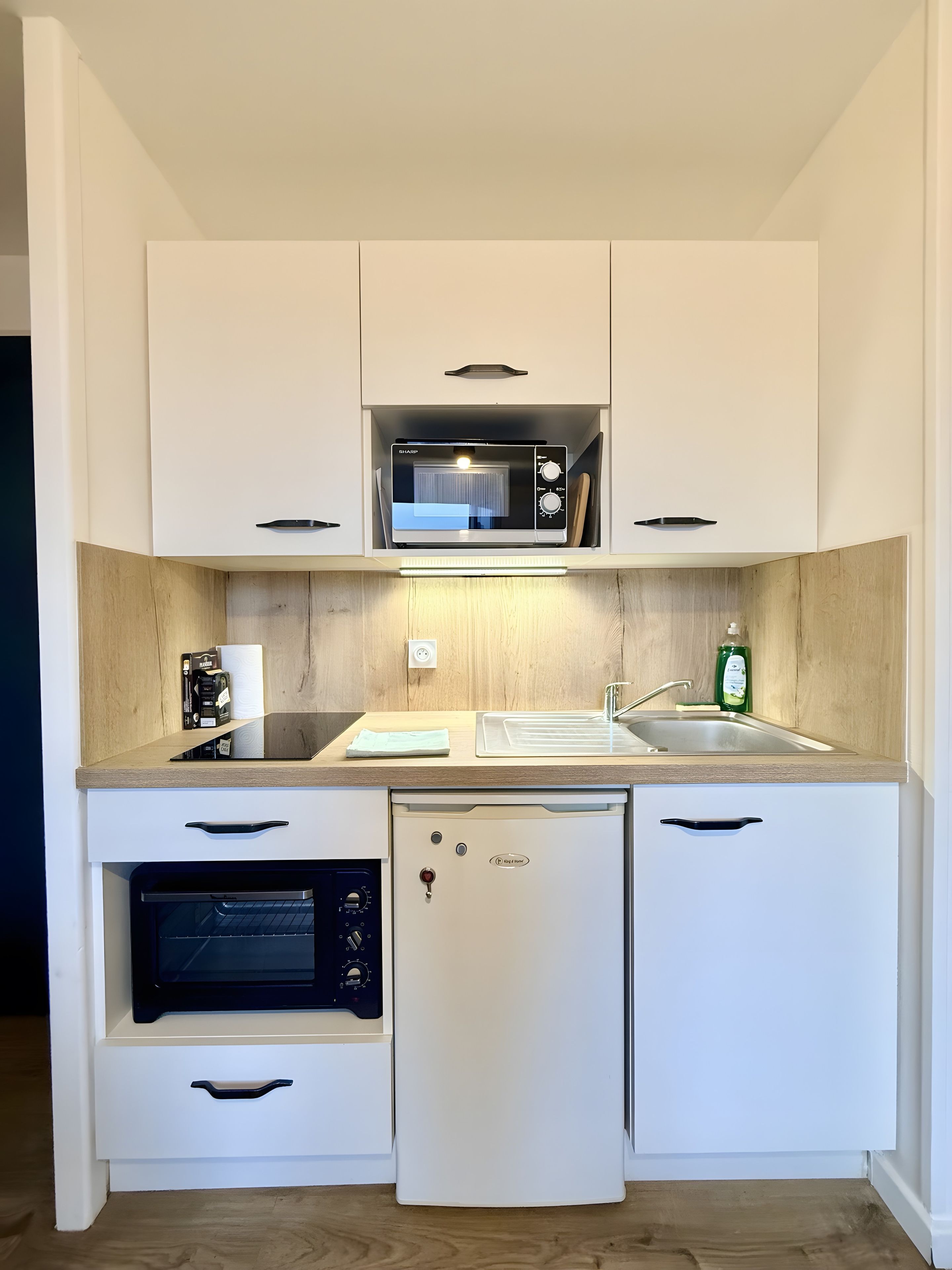 A compact kitchen with a microwave, oven, and refrigerator, featuring white cabinetry and a wooden countertop.