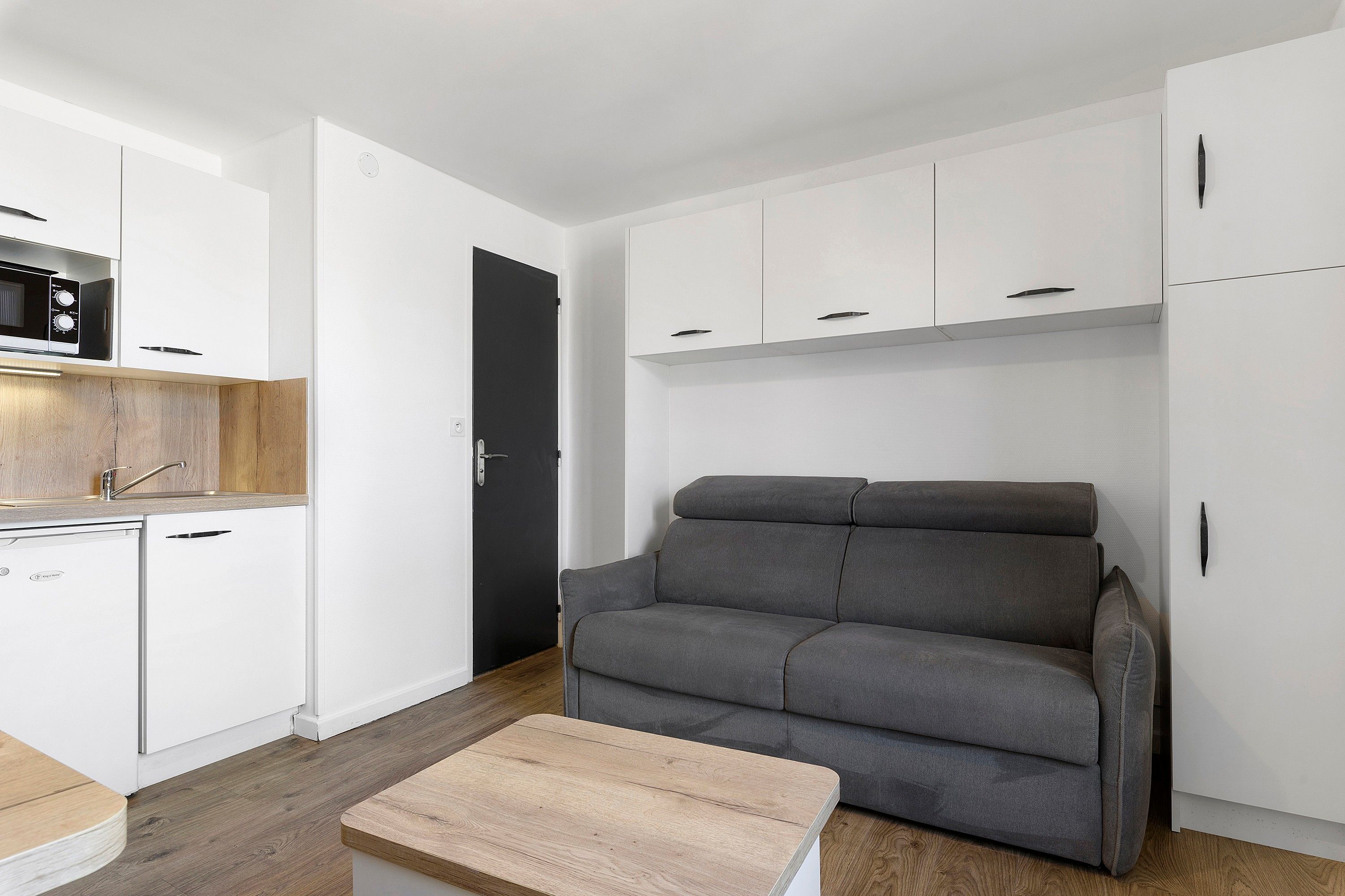 A compact living space with a kitchenette, a grey sofa, and a small wooden table.