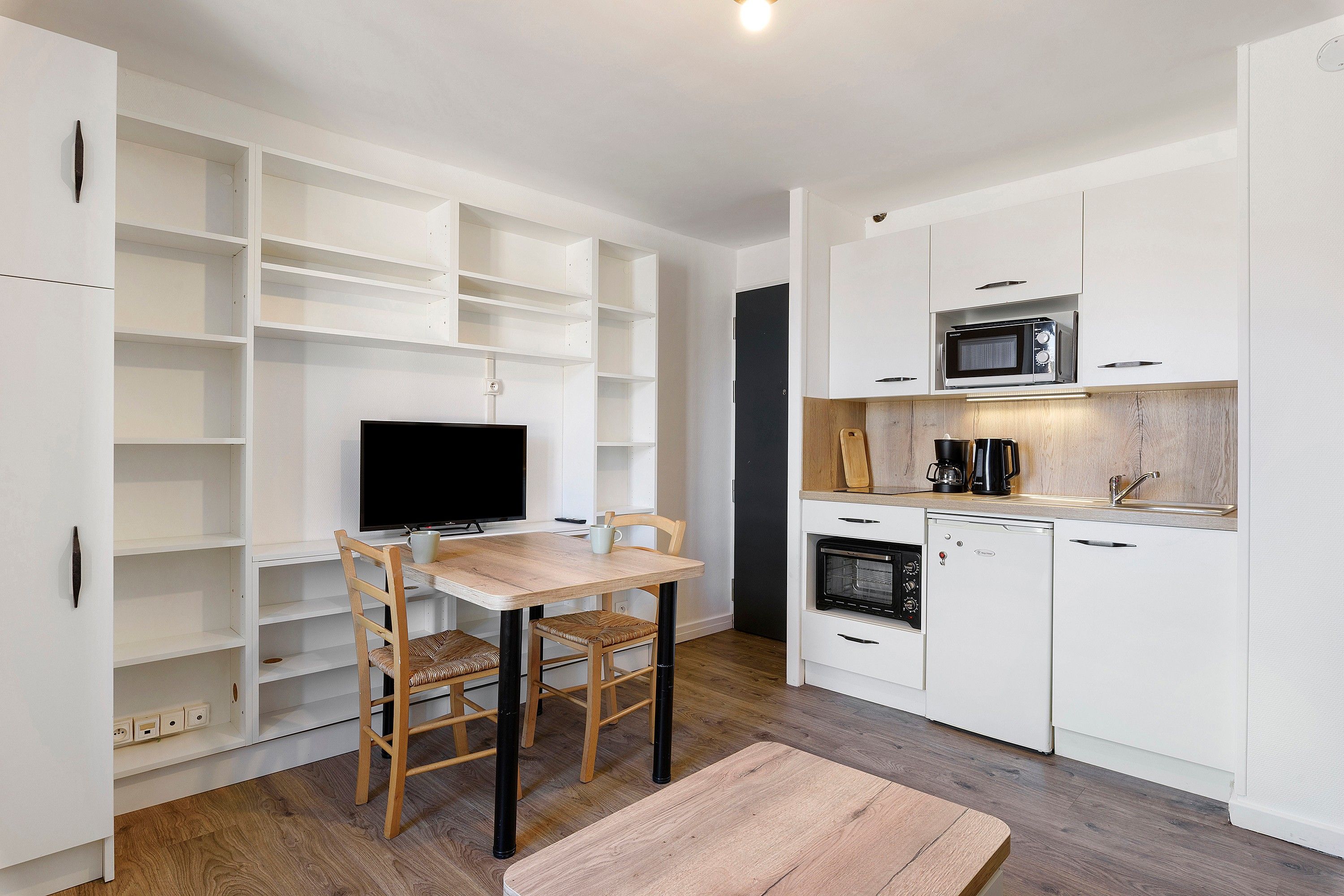 A compact kitchen with white cabinetry, a built-in microwave, and a dining area with a wooden table and chairs.