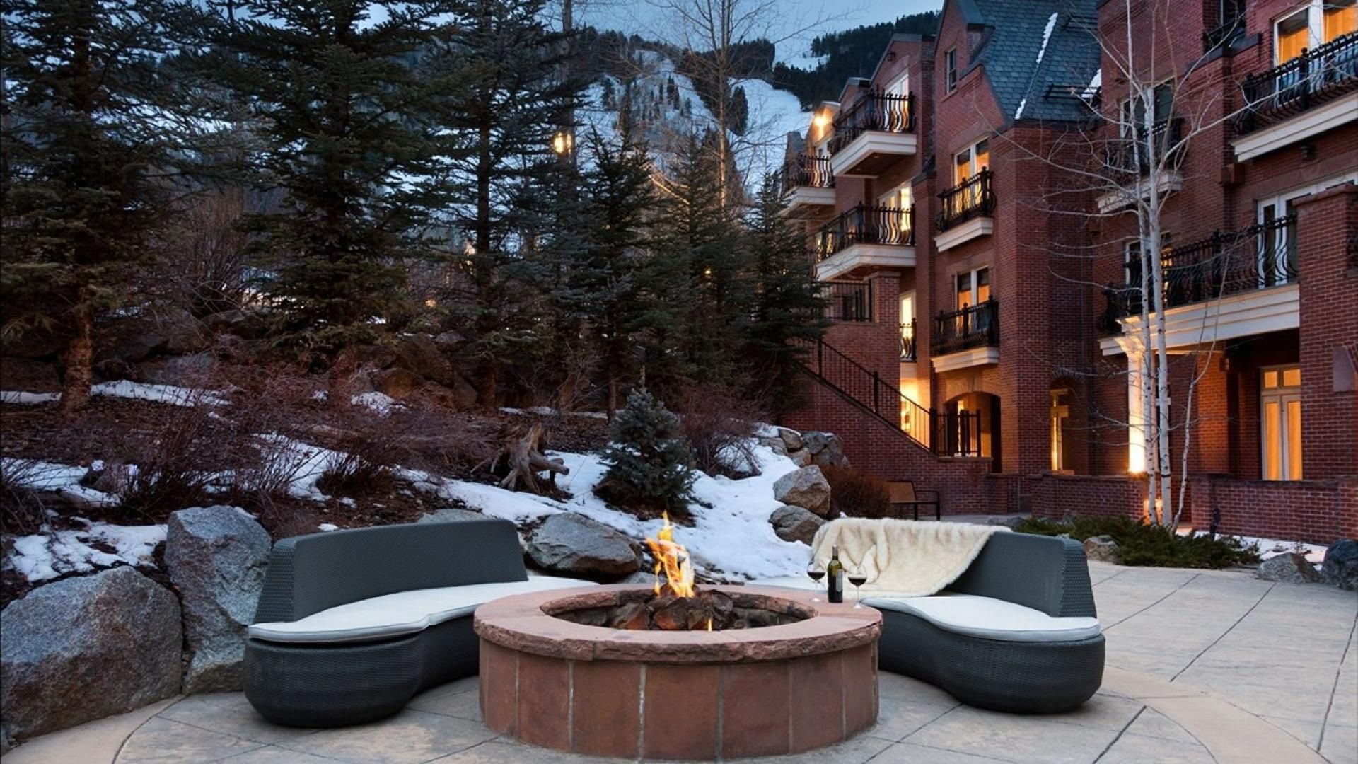 A fire pit with seating, surrounded by snow and trees, with a building in the background.