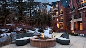 A fire pit with seating, surrounded by snow and trees, with a building in the background.