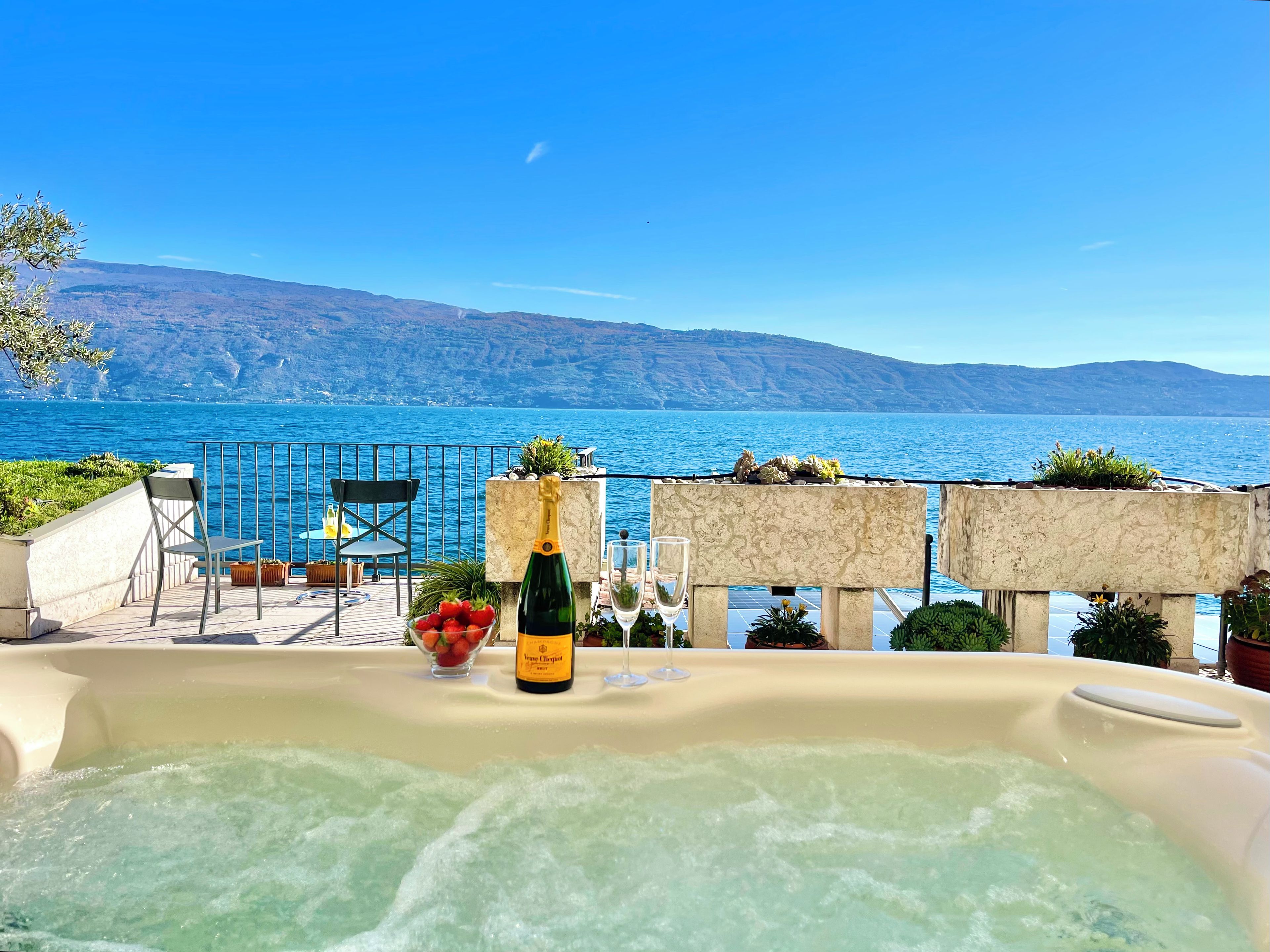 A hot tub with a bottle of champagne and strawberries, overlooking a scenic body of water with mountains in the background.
