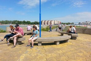 Ein öffentlicher Sitzbereich mit Bänken und einem blauen Pfosten, mit Blick auf einen Strand und das Wasser.