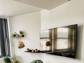 A modern living room with a large flat-screen TV mounted on the wall, a floor lamp, and a small shelf with potted plants. - Mathew Street - City 1 BDR apt sleeps 6 (Merseyside)