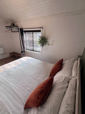 A bedroom with a bed, pillows, a window with blinds, a hanging plant, and a small table. - Stylish and Central - 2 bed, sleeps 4 (Merseyside)