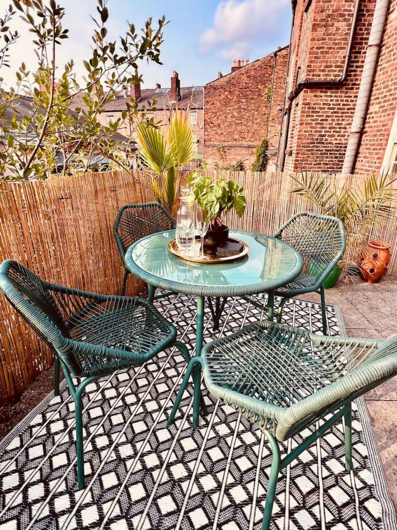 A round glass table with four green wicker chairs, a potted plant, and a bottle of water on the table.