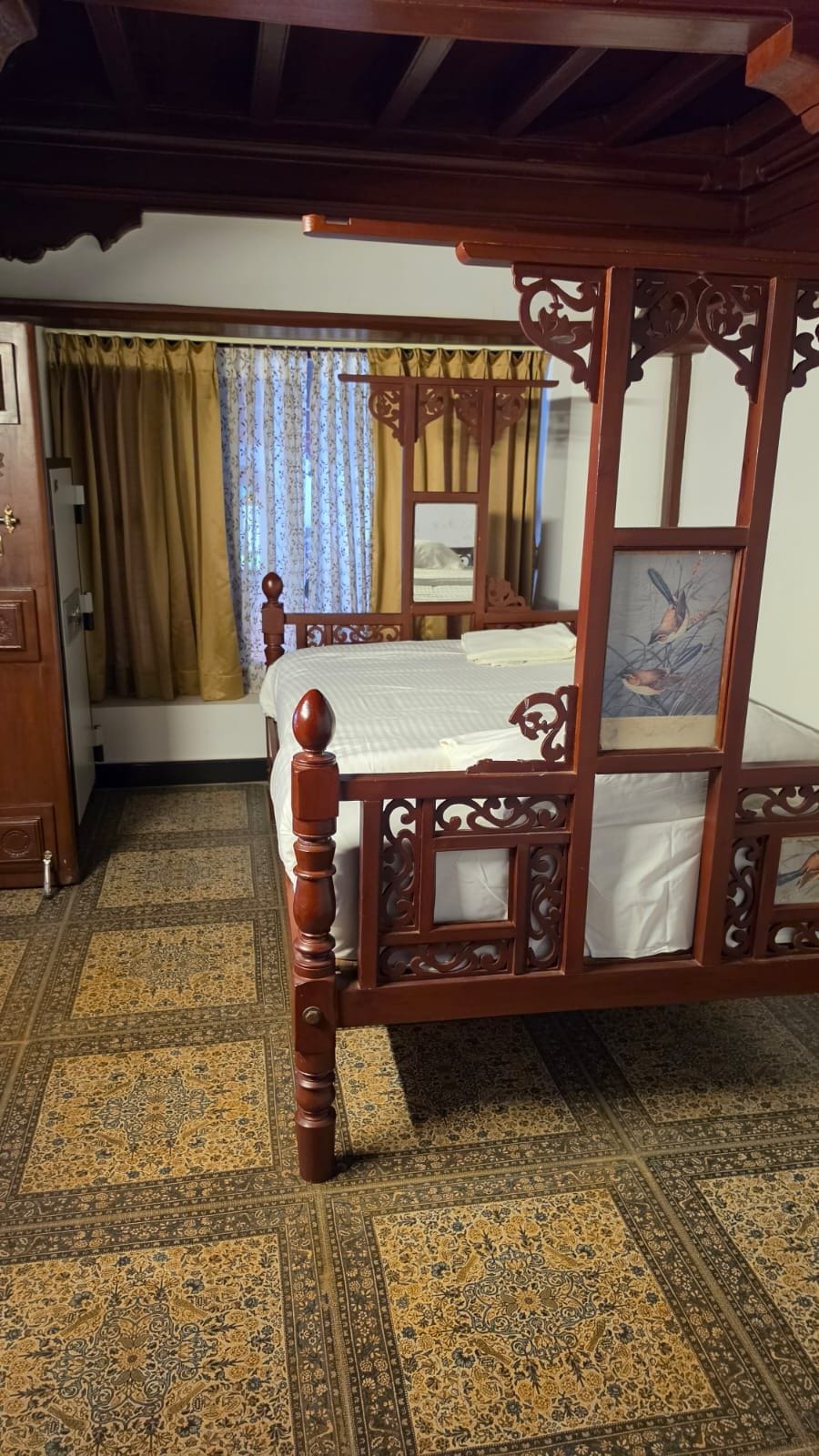 A traditional wooden bed with intricate carvings, a patterned carpet, and a window with curtains.