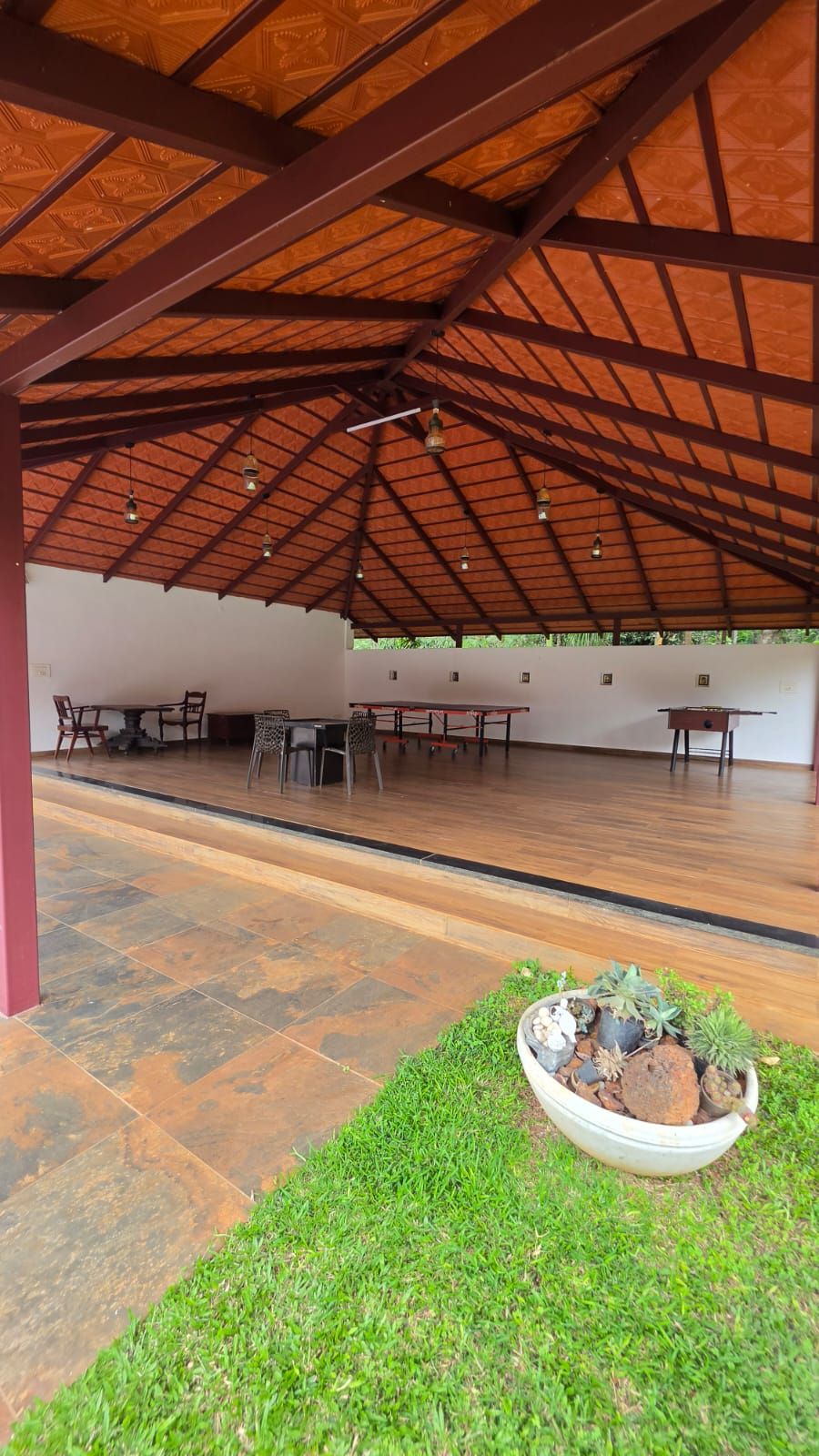 An open-air covered area with tables and chairs, a grassy area with potted plants, and a patterned ceiling.