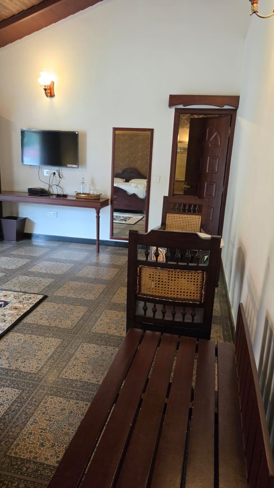 Una habitación con un banco de madera, un televisor fijado en la pared, una cama con cabecero y un espejo.