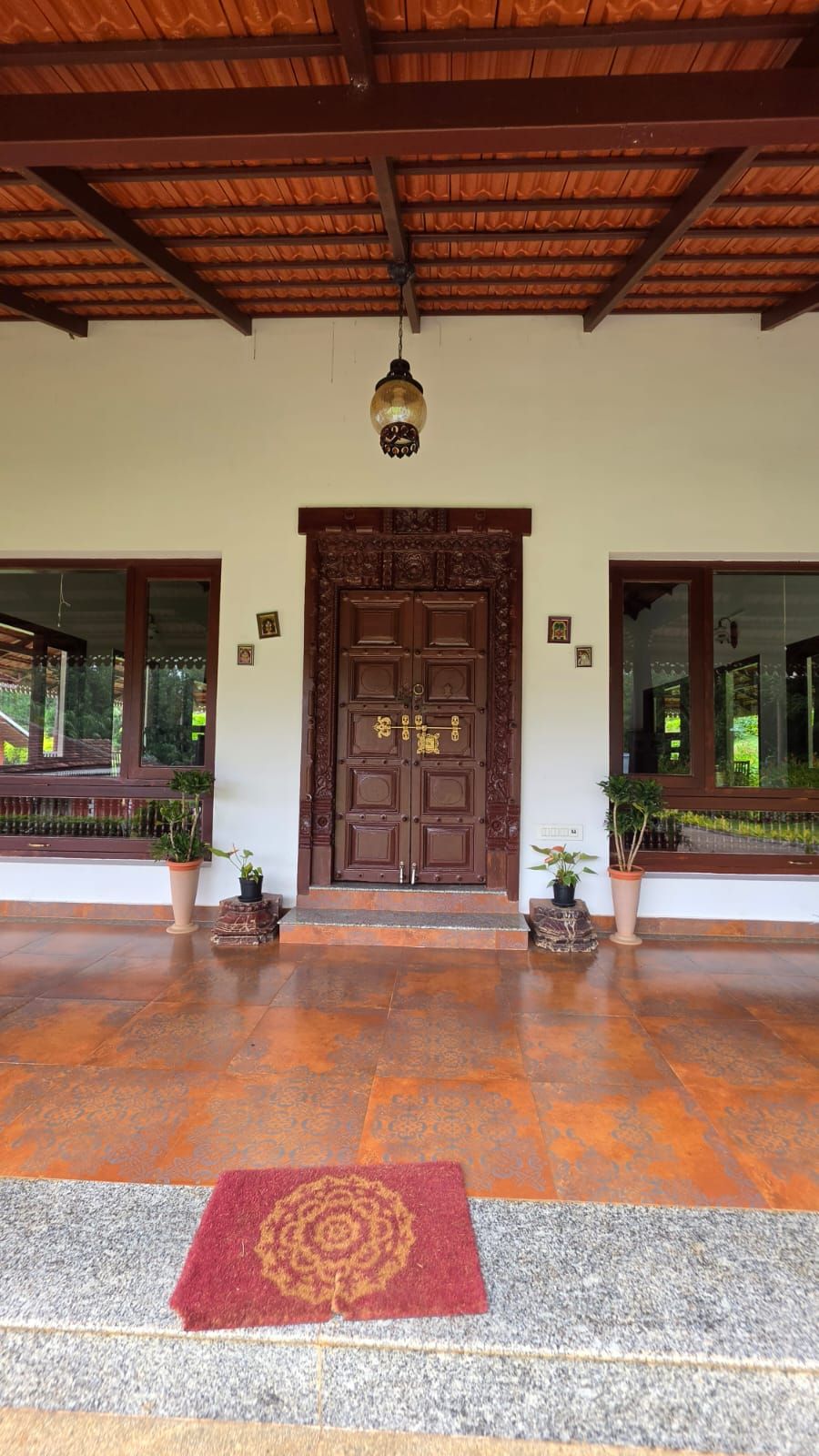 A wooden door with intricate carvings, flanked by potted plants, leading to a covered entrance with a tiled floor.