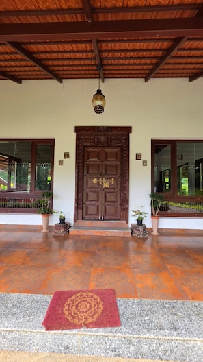 A wooden door with intricate carvings, flanked by potted plants, leading to a covered entrance with a tiled floor.