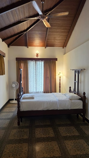 A bedroom with a wooden ceiling, a bed, a standing fan, and a window with curtains.