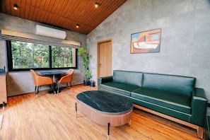 A modern interior with a wooden ceiling, a green sofa, a round coffee table, and a dining area with a table and chairs. - ELIVAAS Signature Nila Forrest Cottage (Mahabalipuram)