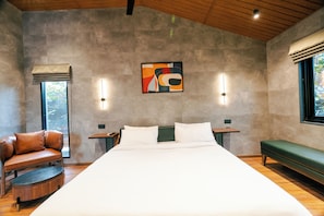 A modern bedroom with a large bed, a green bench, a leather chair, and a wall-mounted artwork. - ELIVAAS Signature Nila Forrest Cottage (Mahabalipuram)