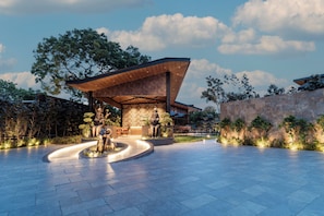 A modern building with a unique roof structure, surrounded by a landscaped area with seating and a water feature. - ELIVAAS Signature Nila Forrest Cottage (Mahabalipuram)