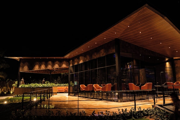 A modern building with a glass facade, outdoor seating area, and a wooden ceiling.