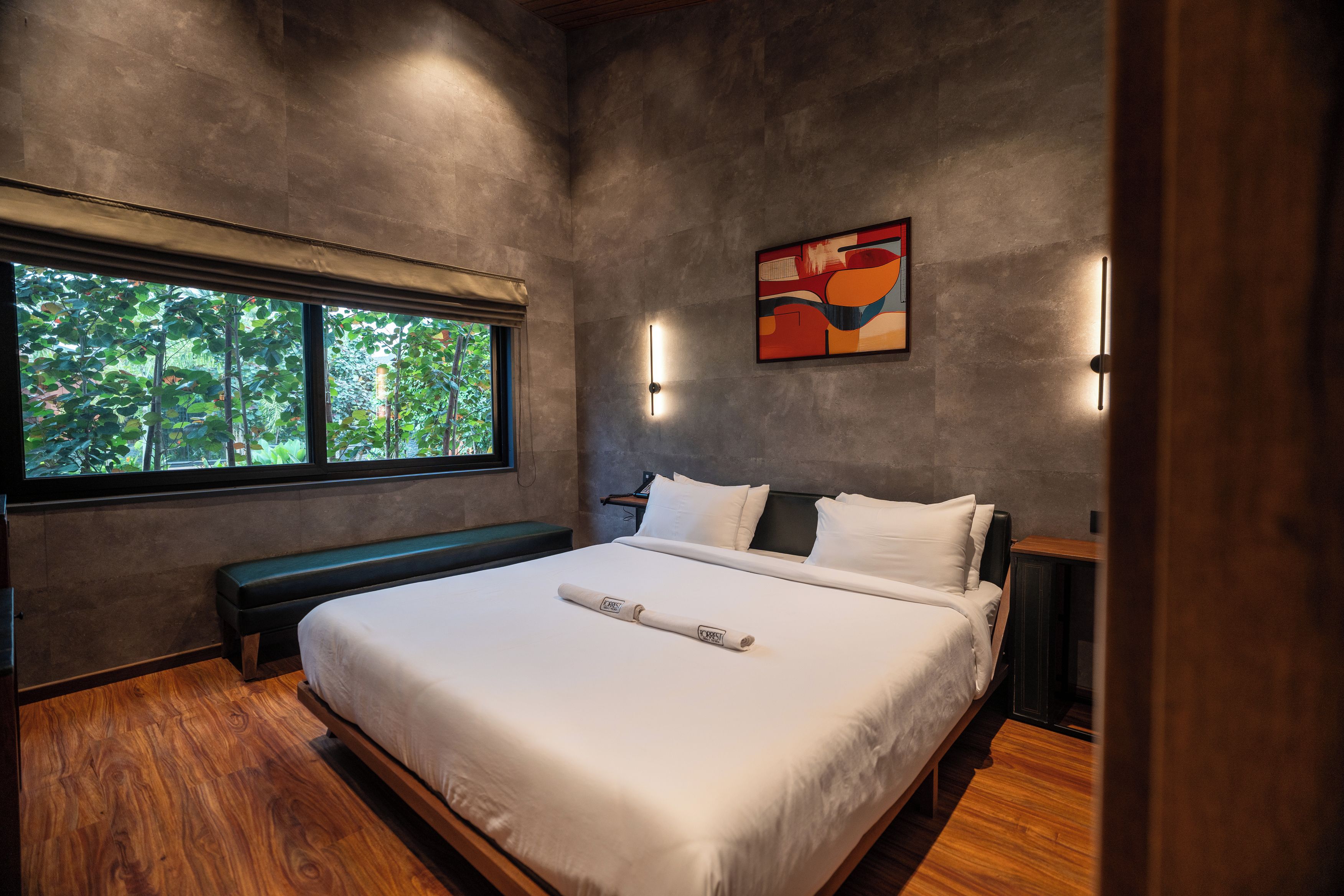 A modern bedroom with a large bed, a bench, a painting, and a view of greenery through a window.