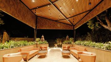 An outdoor seating area with multiple sofas and tables, surrounded by plants and under a wooden roof structure.