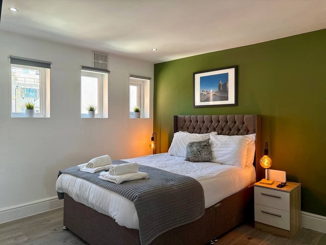 Un dormitorio con cama, mesitas de noche, un cuadro en la pared y ventanas.