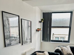 A room with a black sofa, two framed pictures on the wall, a window with curtains, and a small shelf with plants. - Mathew Street - City 1 BDR apt sleeps 6 (Merseyside)