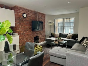 A living room with a brick wall, a flat-screen TV, a sofa, a coffee table, and a dining area with chairs and a glass table. - City centre 2 BDR Sleeps 8 (Merseyside)