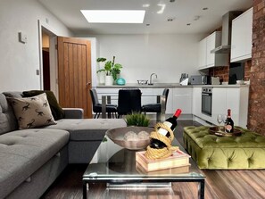 Un soggiorno moderno con un divano, un tavolino in vetro e un poggiapiedi verde. Si intravede in secondo piano una cucina con mobili bianchi e una parete in mattoni.
