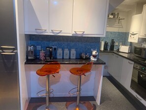 Eine kleine Küche mit weißen Schränken, einer blauen Fliesenspiegel und zwei orangefarbenen Barhocker.