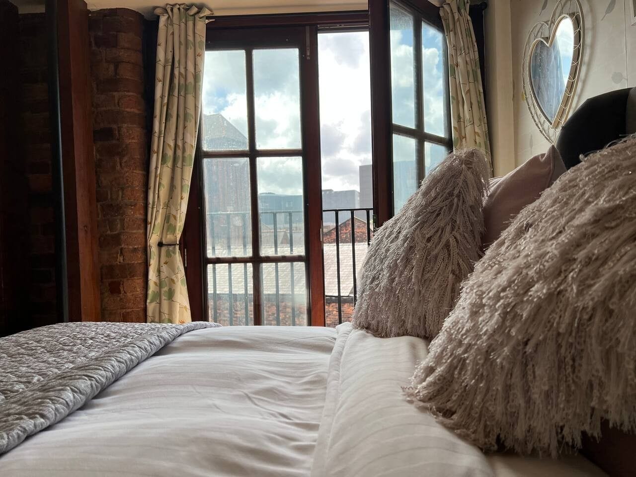 A bed with a white comforter and fluffy pillows, a window with curtains, and a heart-shaped mirror on the wall.