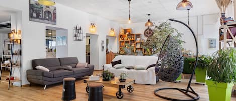 A modern living room with a grey sofa, a coffee table, and a hanging chair. There are plants, a bookshelf, and a large window.