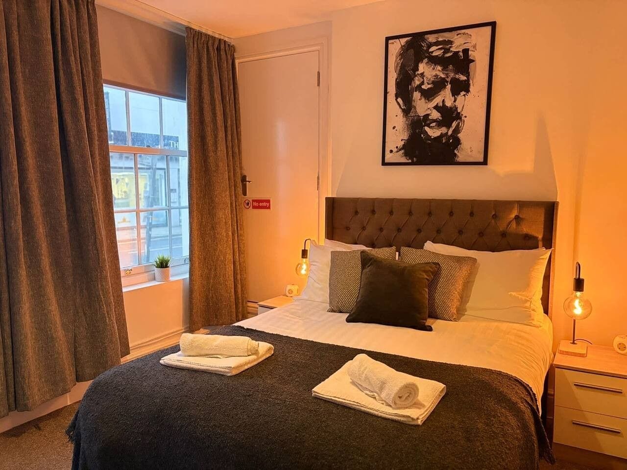 Una habitación de hotel con una cama, dos mesitas de noche, un ventanal con cortinas, una obra de arte colgada en la pared y un cartel de 'No admitido'.