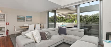 A modern living room with a sectional sofa, a dining area with a table and chairs, and large sliding glass doors leading to a balcony with greenery.